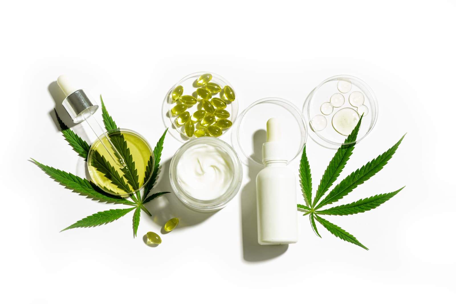 A flat-lay photograph of various cannabis products on a white background, including oil dropper bottles, a jar of topical cream, and several capsules in petri dishes. Two fresh green cannabis leaves are tucked under the glass containers.